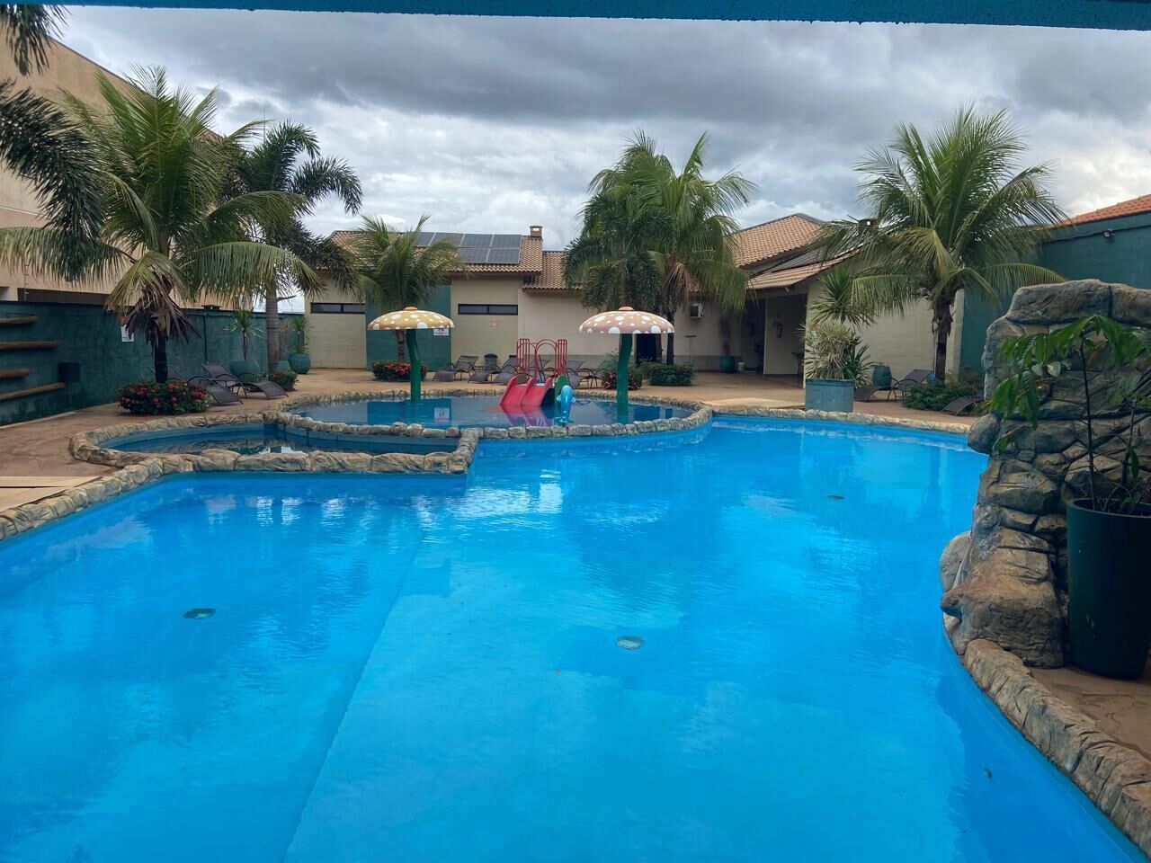 A swimming pool with a stone border, palm trees, and a cloudy sky.