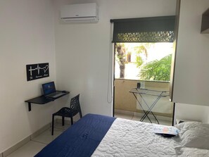 A bedroom with a bed, a desk with a laptop, a chair, an air conditioner, and a view of a balcony with plants.