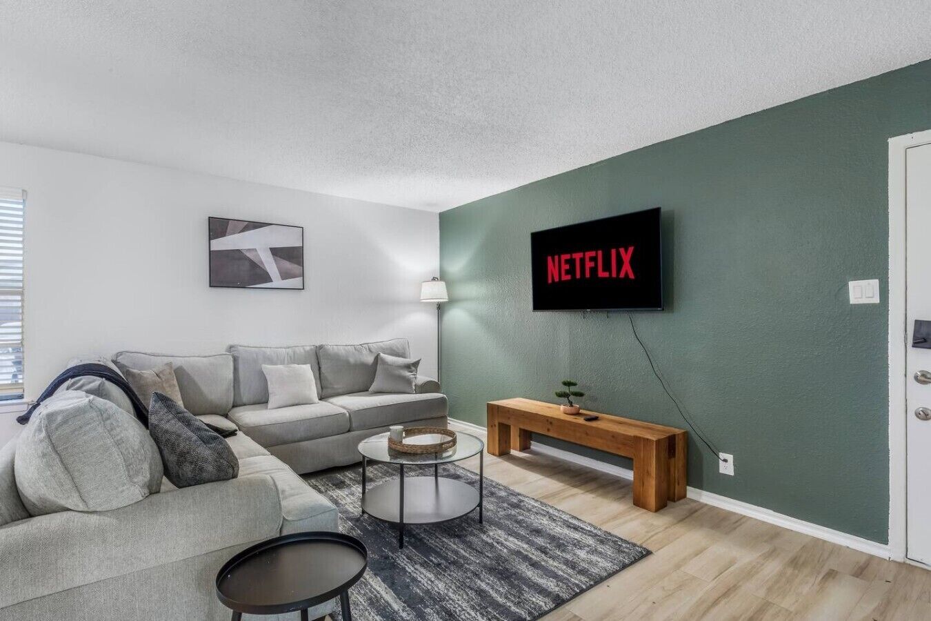 Ein modernes Wohnzimmer mit einem grauen Sofa, einem Couchtisch, einem an einer grünen Wand montierten Fernseher und einer hölzernen Bank.