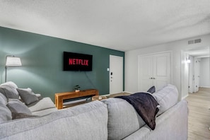 Ein Wohnzimmer mit Fernseher, der 'NETFLIX' anzeigt, einem Sofa, einer Bank und einer Lampe.