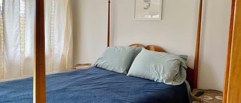A bedroom with a four-poster bed, a bedside table, a framed artwork on the wall, and a window with curtains.