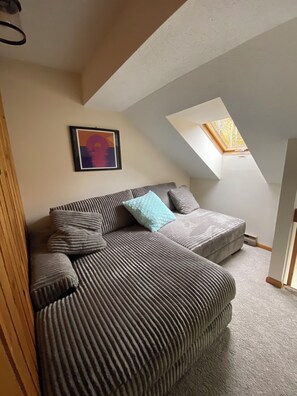 A loft-style room with a sloped ceiling, a skylight, a framed abstract painting, and a large sofa with multiple cushions. - Where the lakes meet the mountains. (Thornton)