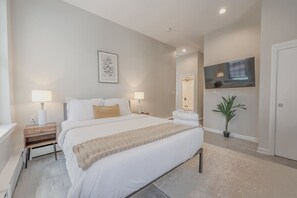 A bedroom with a bed, bedside tables, a TV, a plant, and a door. - Back Bay Area Elegant Studio Apt (Boston)