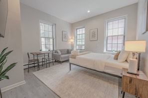 A modern bedroom with a bed, bedside table, lamp, and a small desk with a chair. - Back Bay Area Elegant Studio Apt (Boston)