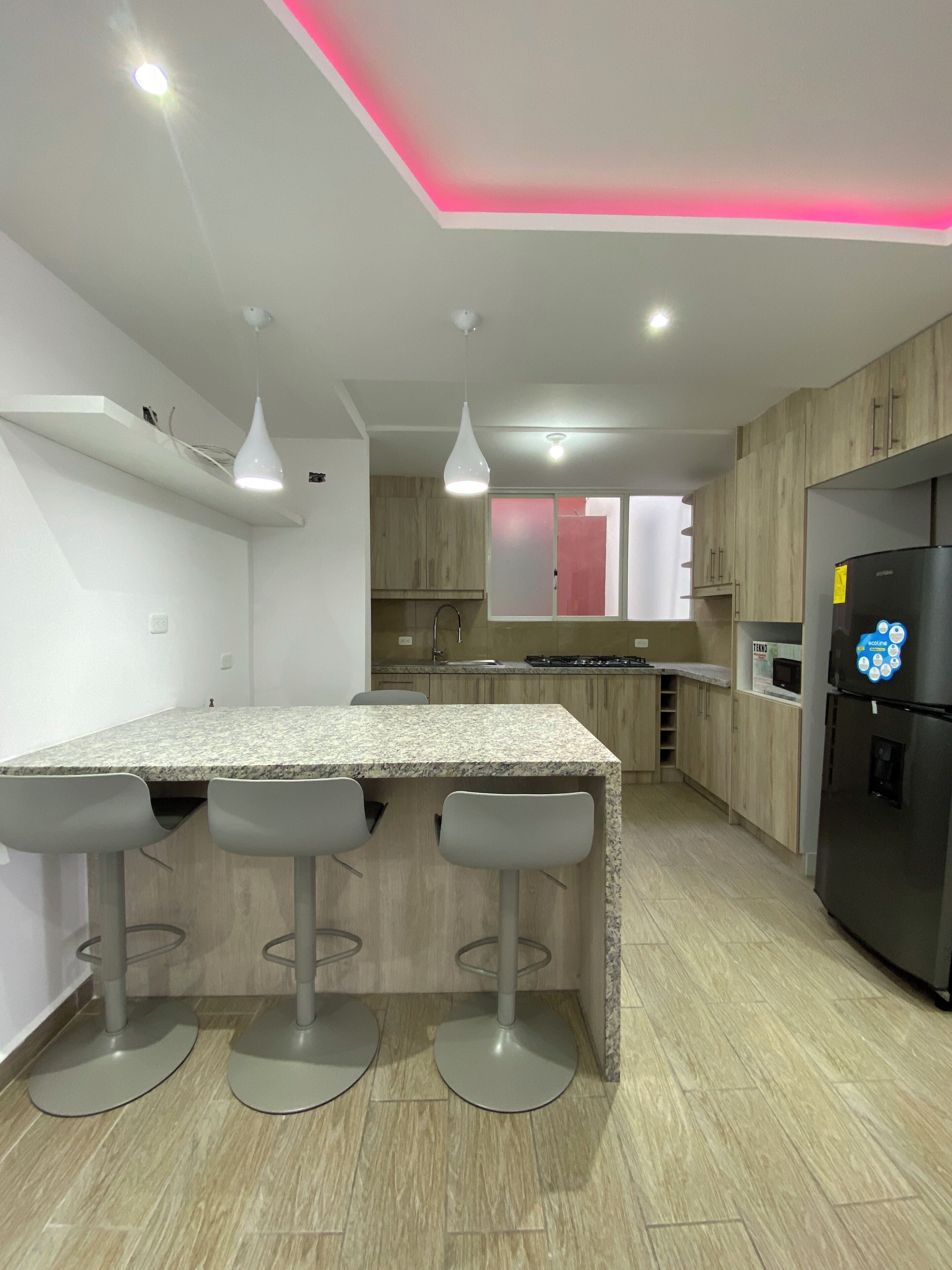 A modern kitchen with a granite countertop, bar stools, a refrigerator, and wooden cabinets.