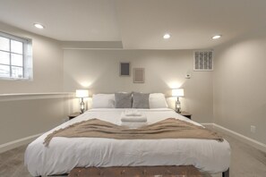A bedroom with a large bed, two bedside lamps, a window with blinds, and a ceiling vent. - Spacious & Modern 1 Bedroom | Back Bay (Boston)