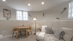 A cozy living room with a round wooden table, a sofa with cushions, a floor lamp, and wall art. - Spacious & Modern 1 Bedroom | Back Bay (Boston)
