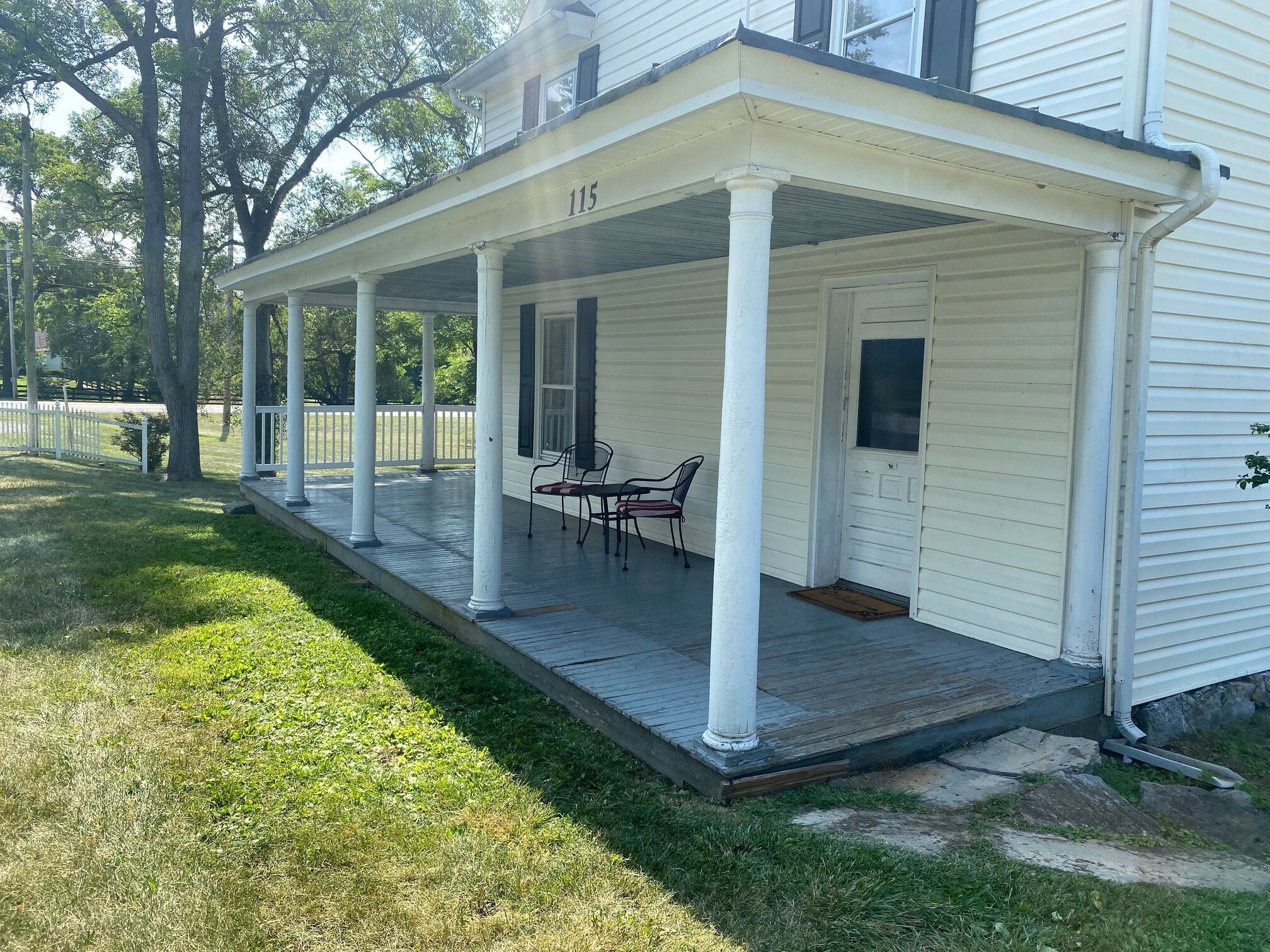 A white house with a covered porch, numbered 115, featuring a small table and chairs.