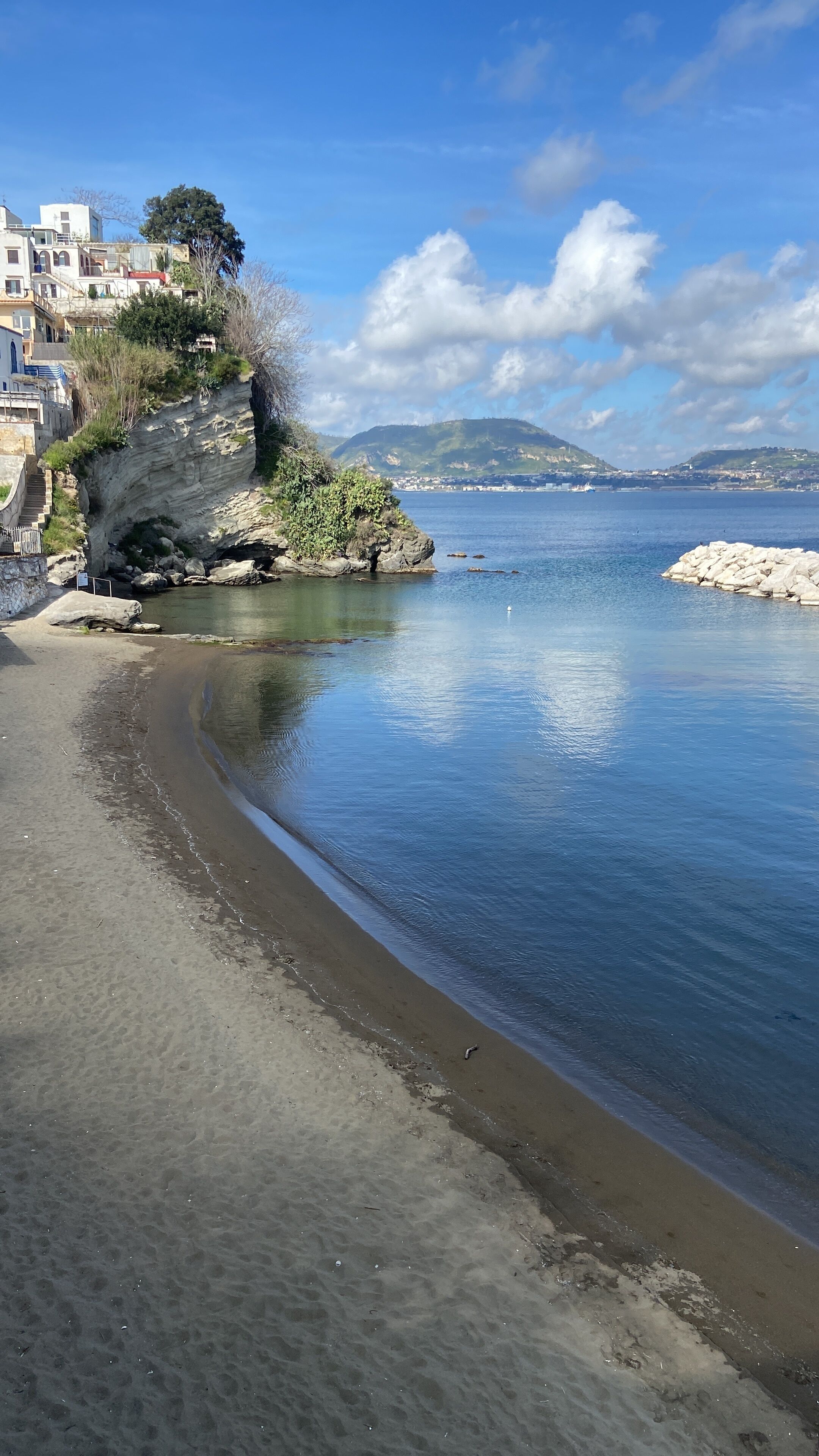 A coastal beach with a rocky shoreline, clear blue water, and a backdrop of buildings and hills.