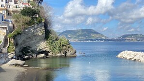 A coastal beach with a rocky shoreline, clear blue water, and a backdrop of buildings and hills. - Vrbo Property (Bacoli)
