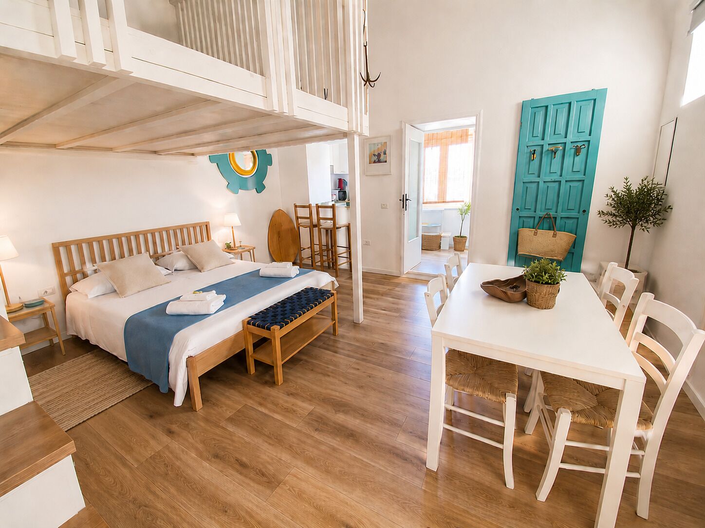 A bedroom with a bed, bedside tables, a bench, a dining table with chairs, and a blue door.