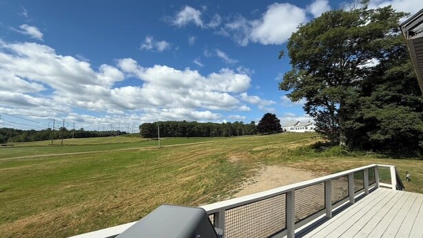 A deck with a grill, overlooking a grassy field and a building in the distance.
