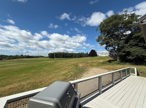 A deck with a grill, overlooking a grassy field and a building in the distance.