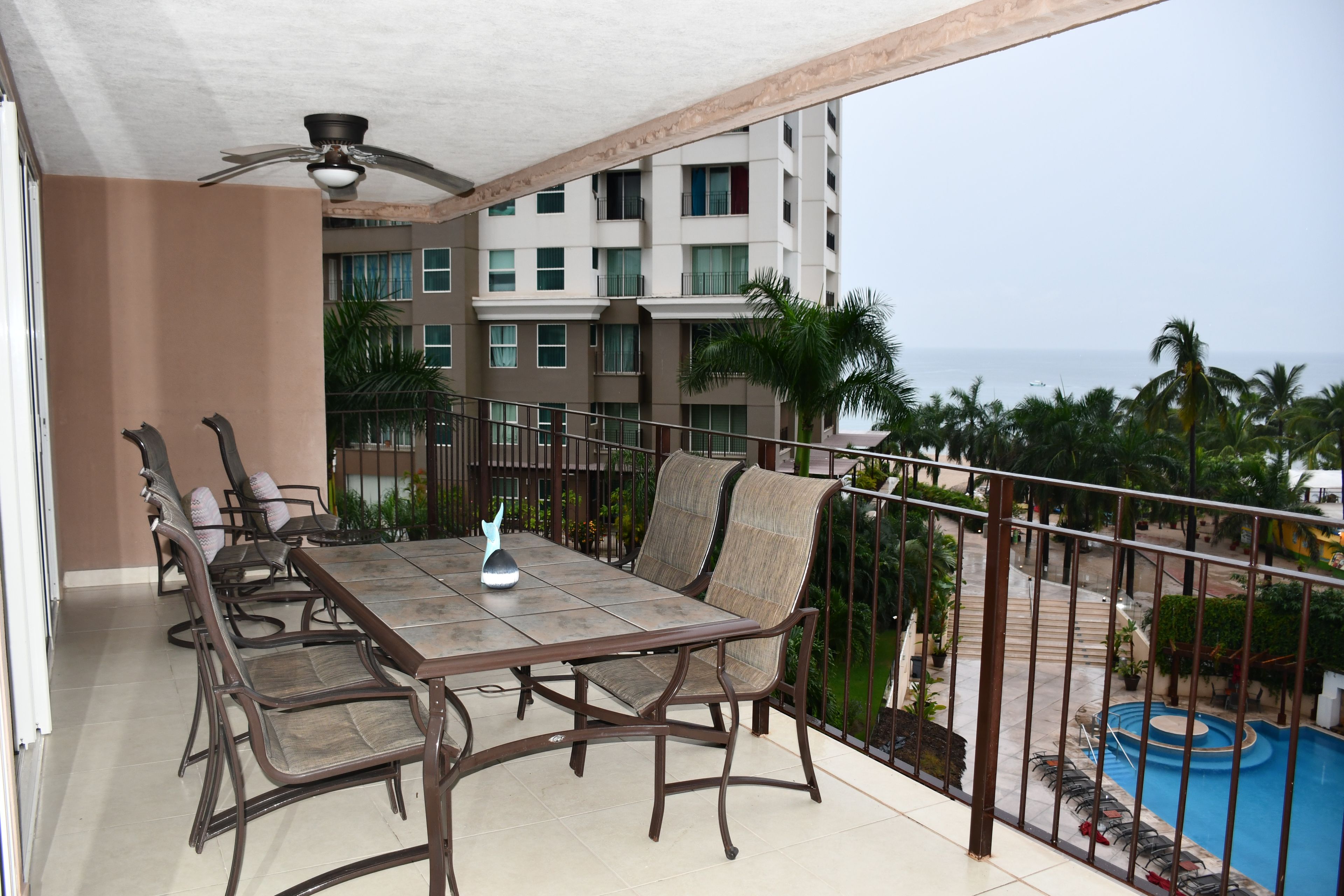 A balcony with a table and chairs, a ceiling fan, and a view of a building and a swimming pool.
