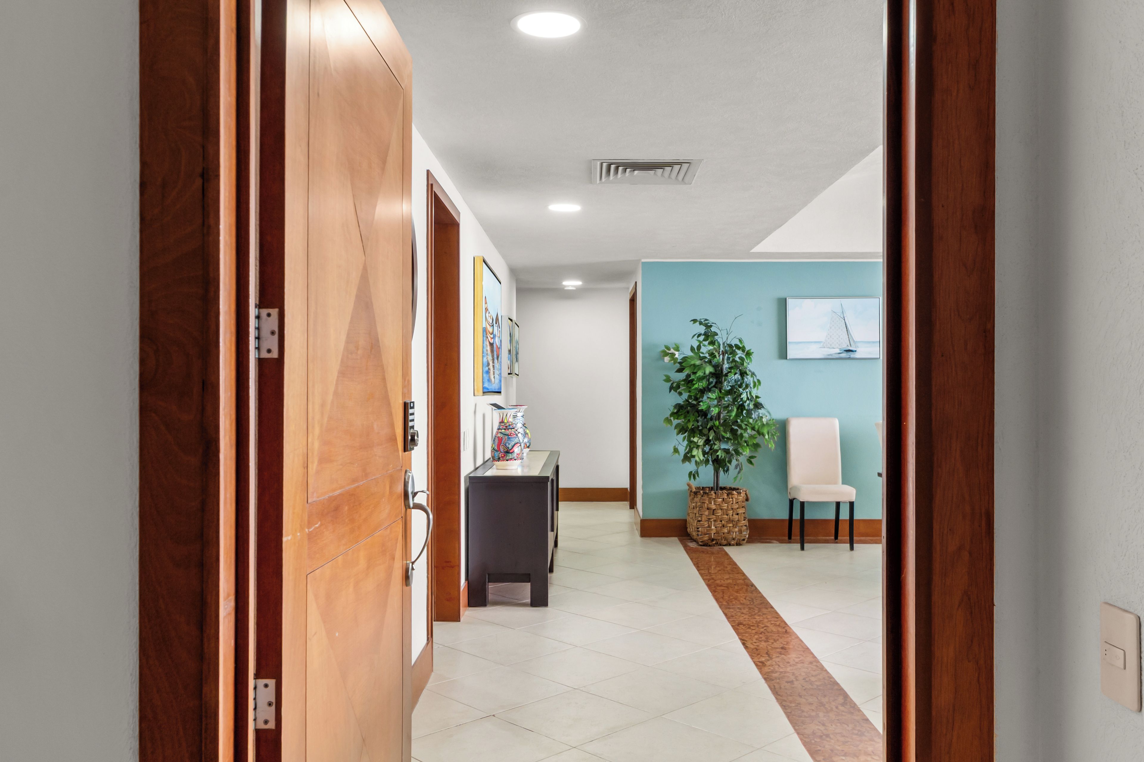 A hallway with a wooden door, a desk with a vase, a chair, and a wall with framed pictures.