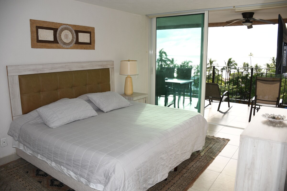 A bedroom with a bed, a nightstand, a lamp, and a view of a balcony with outdoor furniture.