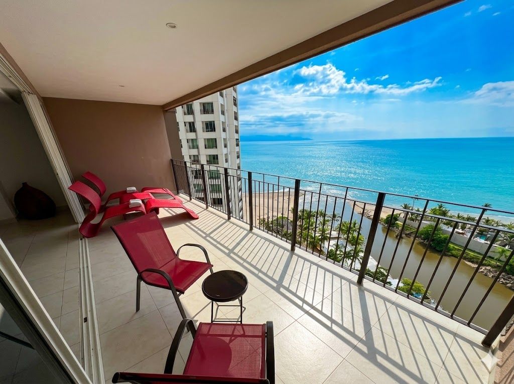 A balcony with red chairs and a table, overlooking a beach and ocean.
