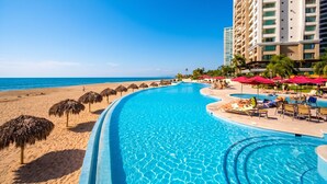 Piscina de un resort con vista a la playa, palmeras y un edificio de gran altura.