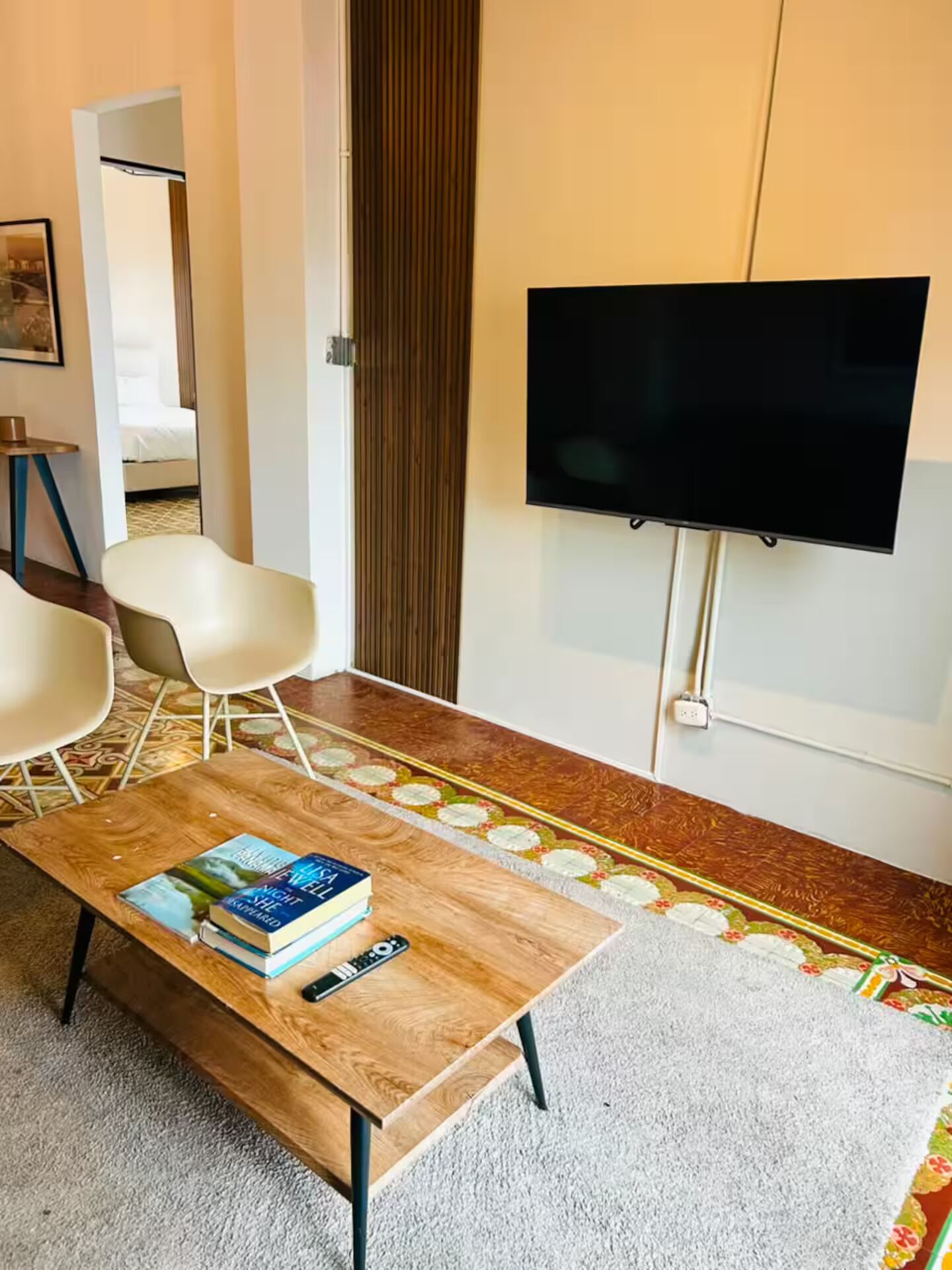 Una sala de estar moderna con un televisor de pantalla plana, una mesa de centro con libros y un control remoto, dos sillas blancas y una alfombra estampada.