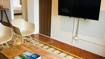 Una sala de estar moderna con un televisor de pantalla plana, una mesa de centro con libros y un control remoto, dos sillas blancas y una alfombra estampada.