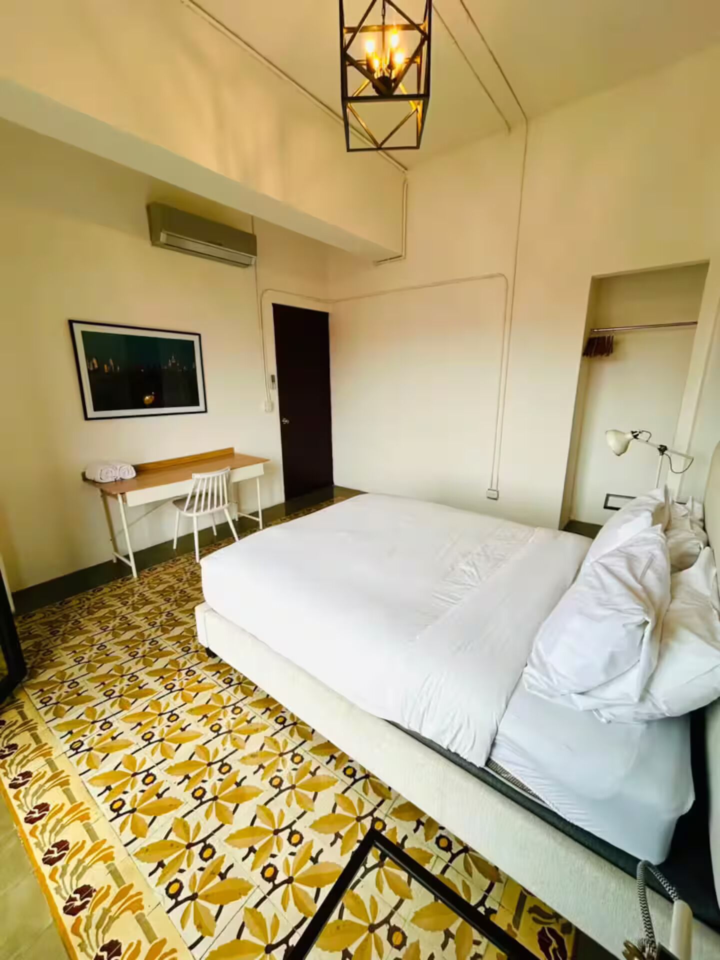 A hotel room with a bed, a desk, a chair, a painting, and a patterned floor.