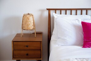 A wooden nightstand with a woven basket and a drawer, next to a bed with white bedding and a pink pillow. - Modern Mesa Retreat Pool Near Gilbert & US-60 (Mesa)