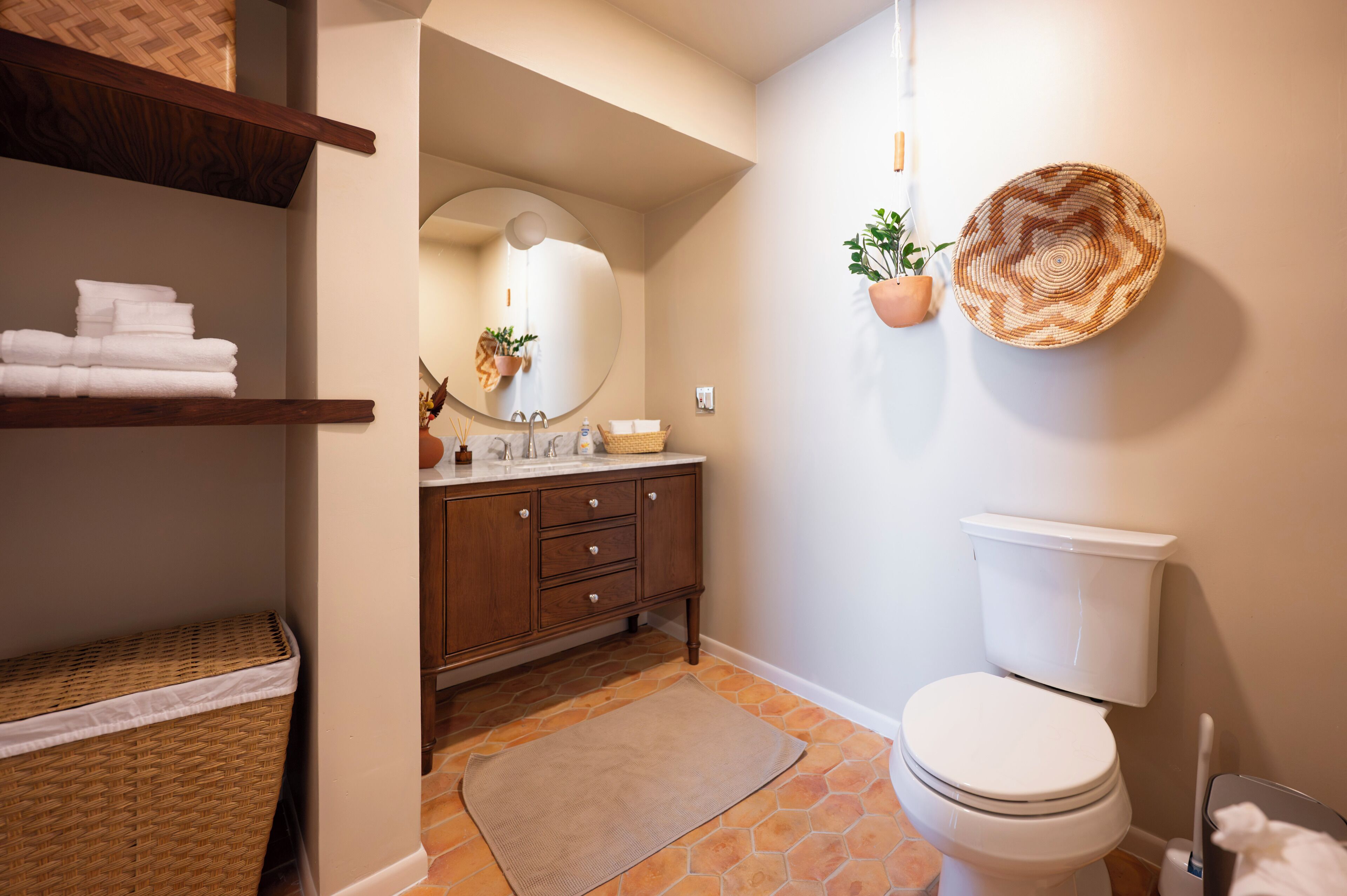 A bathroom with a toilet, a sink with a mirror, a wooden vanity, and a shelf with towels.