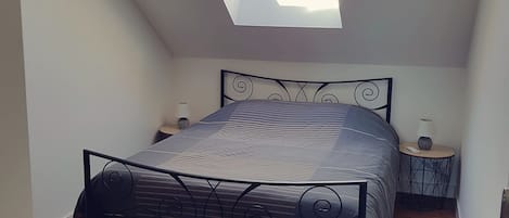 Une chambre avec un plafond en pente, un lit avec un cadre en métal, une petite table de chevet et un puits de lumière.