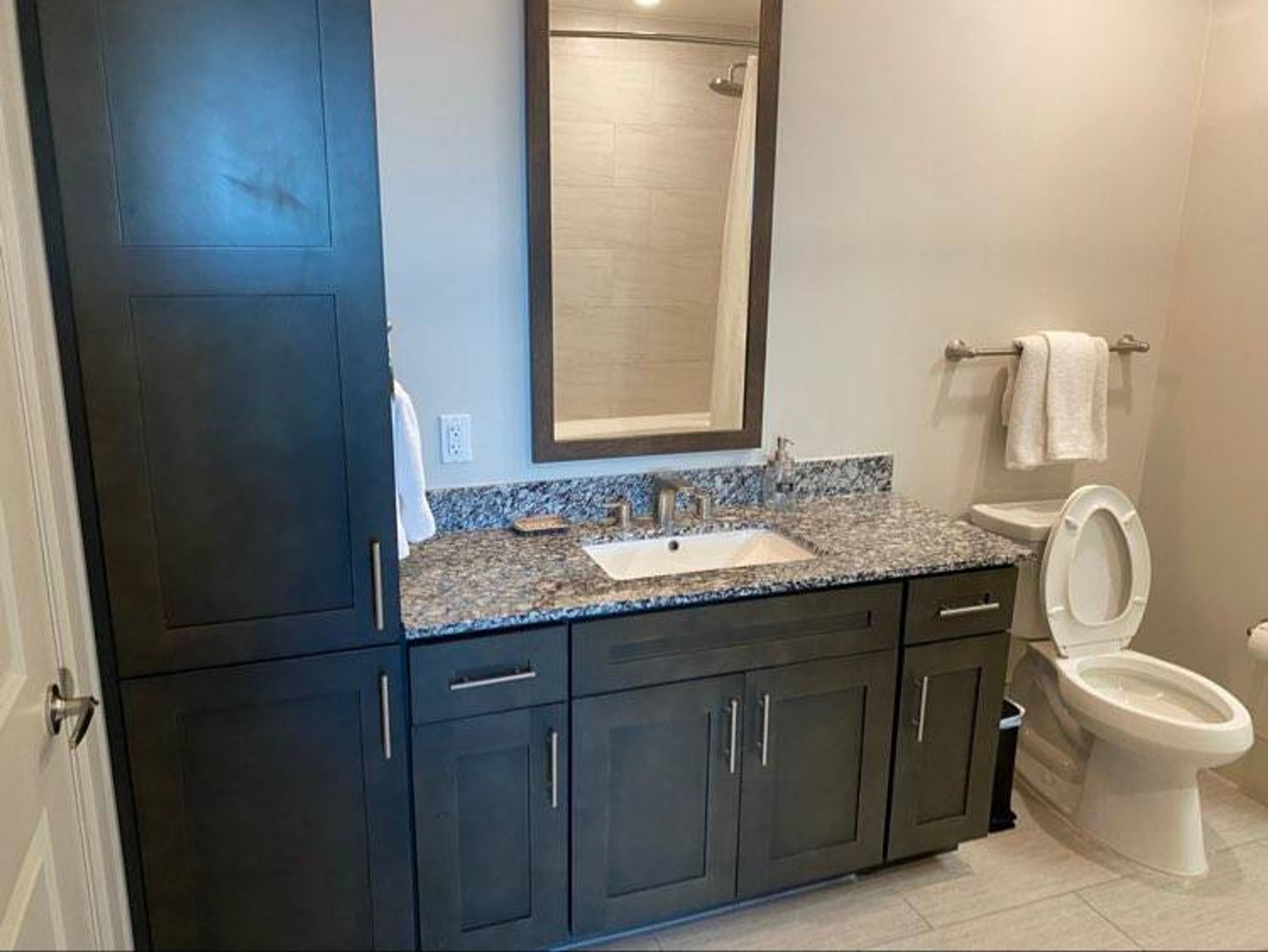 Un baño con muebles oscuros, encimera de granito, lavabo, espejo, inodoro y ducha.