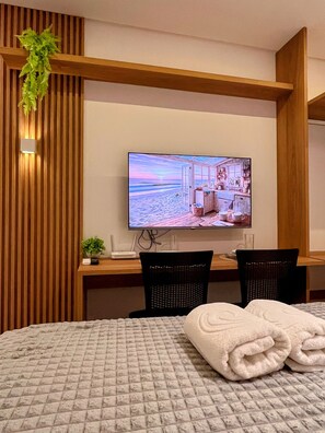Habitación de hotel con una cama, un escritorio con una computadora, dos sillas, un televisor mostrando una playa y una planta en maceta.