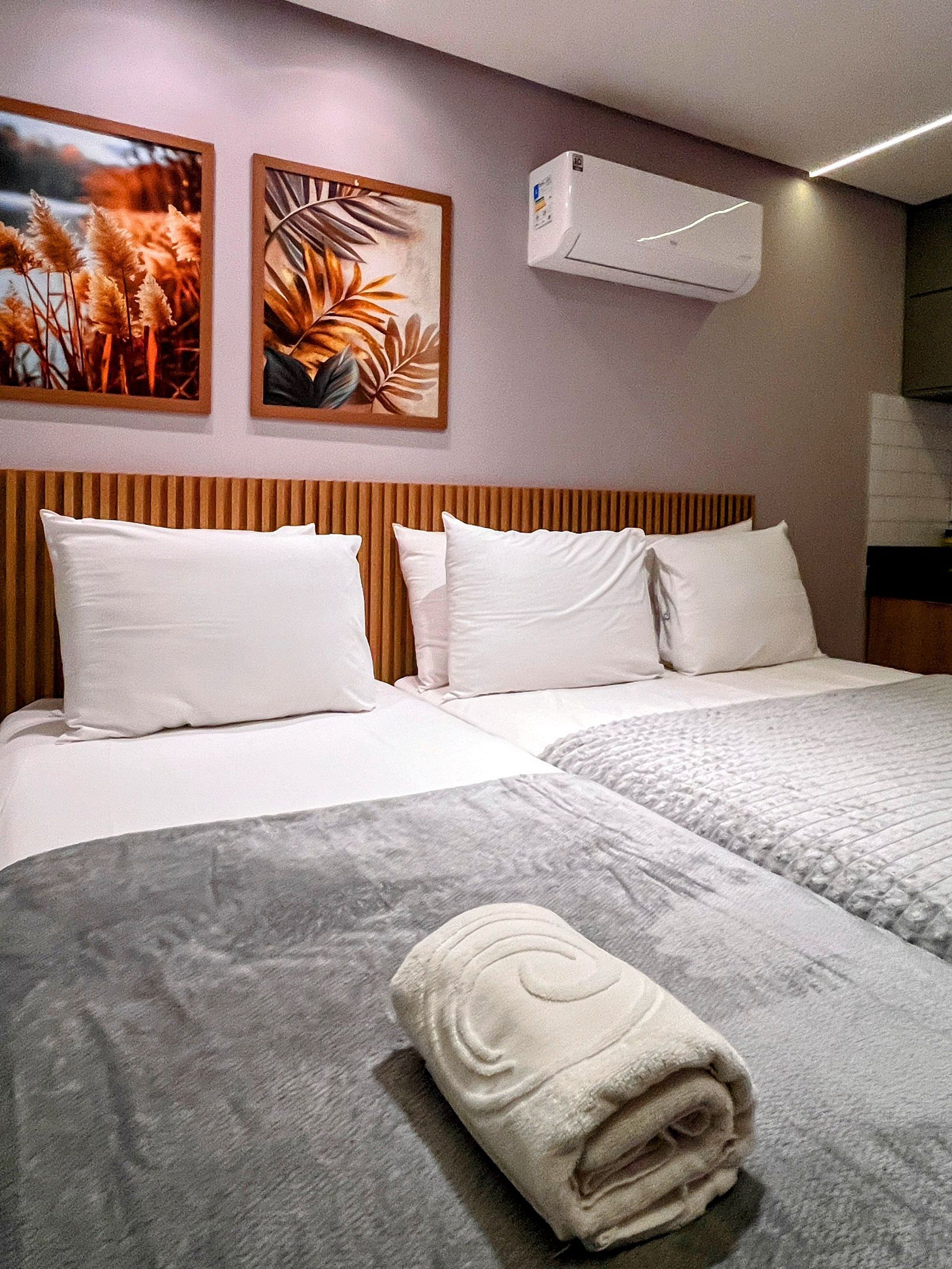 A hotel room with two beds, a wooden headboard, white bedding, a rolled towel on the bed, and two wall-mounted air conditioning units.