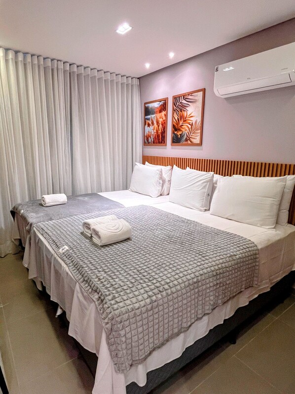 A hotel room with a large bed, two bedside tables, a flat-screen TV, a wall-mounted air conditioner, and a window with curtains.