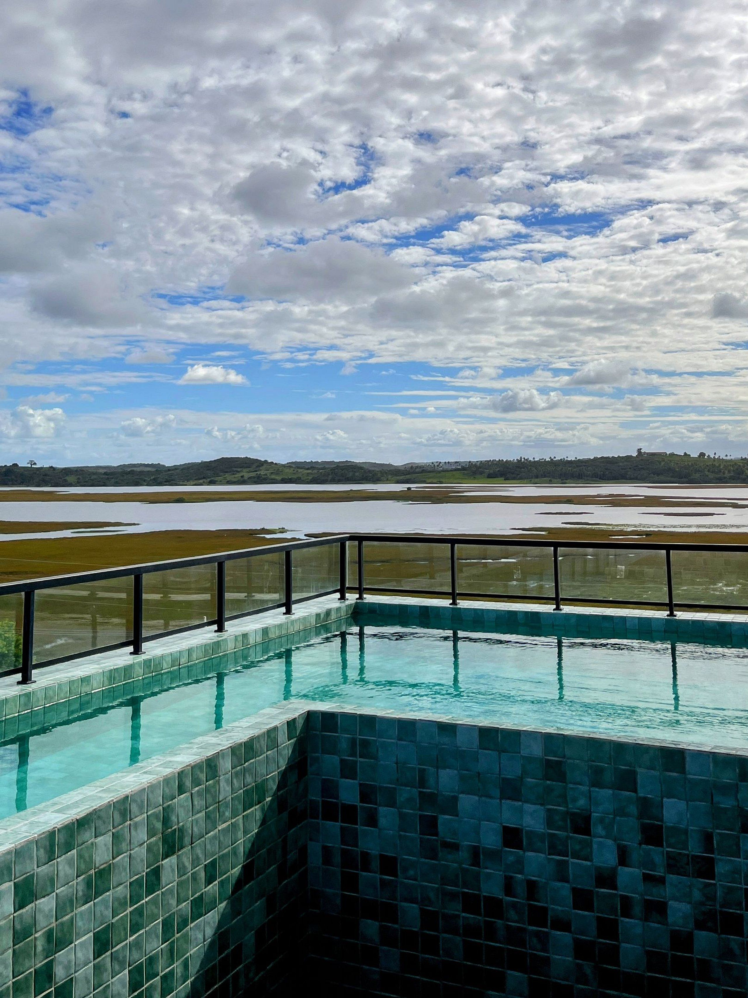 Une piscine dont le bord est recouvert de carreaux, entourée d’une rambarde en métal, et qui offre une vue sur un paysage plat avec des collines lointaines sous un ciel partiellement nuageux.