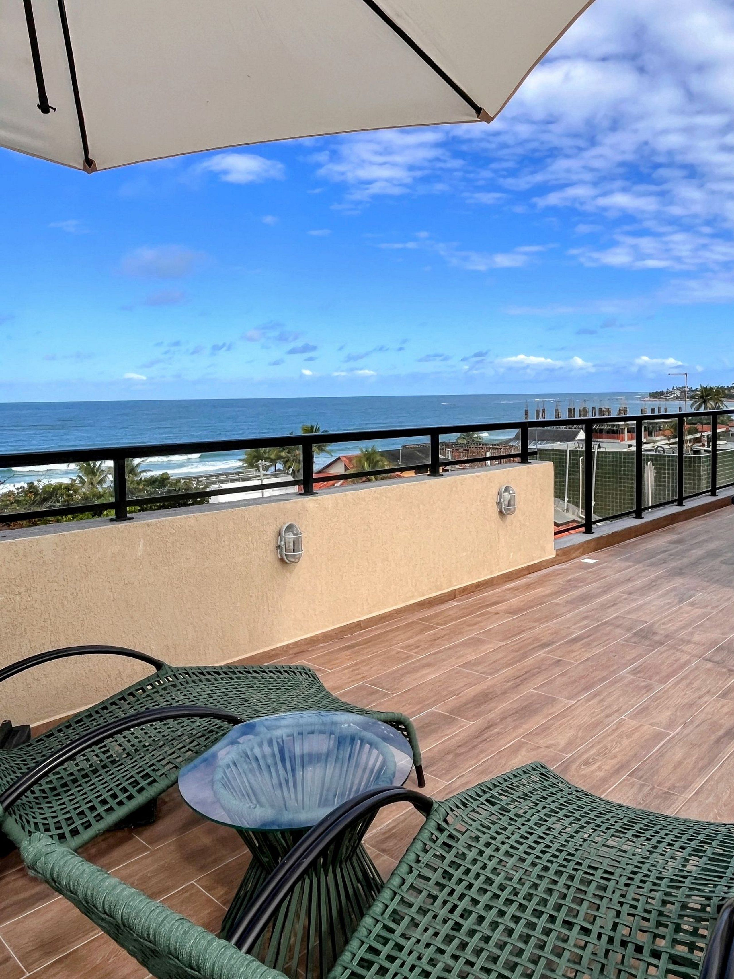 A balcony with a glass table and chairs, overlooking a beach and ocean.