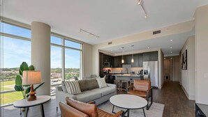 A modern living room with a sofa, coffee table, and chairs, a kitchen with dark cabinets and stainless steel appliances, and large windows offering a view of the outdoors. - Preston Royal Double I Premium 2BR in Dallas (Dallas)