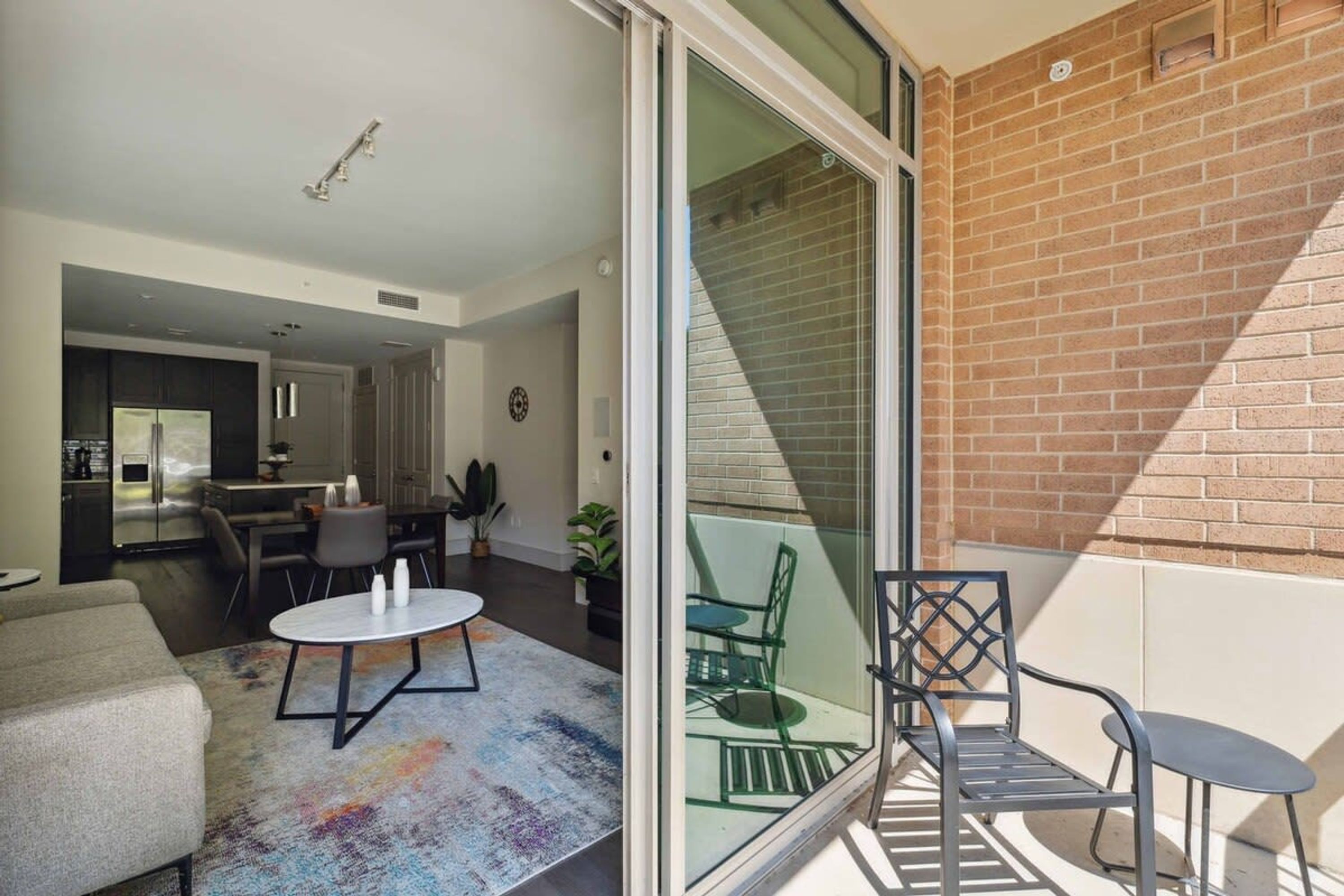 A modern living room with a sofa, a round table, and a glass door leading to a balcony with a brick wall and a chair.