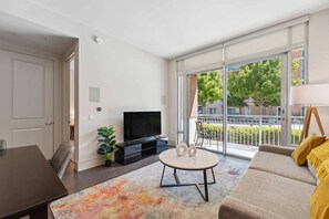 A modern living room with a sofa, a TV, a dining table, and a large window with a view of the outdoors. - Preston Douglas Single I Premium 1BR in Dallas (Dallas)