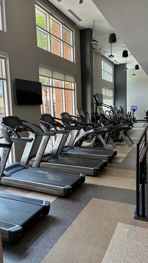 A gym with multiple treadmills lined up against a wall with windows, a flat-screen TV mounted on the wall, and ceiling lights. - Preston Douglas Double I Premium 2BR in Dallas (Dallas)
