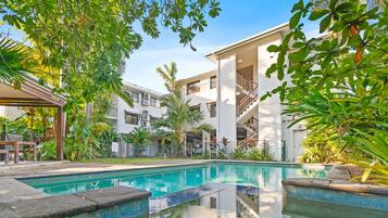 Un área de piscina con una piscina azul cristalina, rodeada de exuberante vegetación y un edificio de varios pisos con balcones.