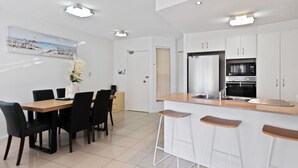 A modern kitchen with white cabinets, a built-in oven, and a stainless steel sink. There is a dining area with a wooden table and black chairs. - Cairns Hideaway - Pool and Spacious (Cairns)