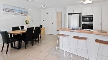 Una cocina moderna con armarios blancos, horno integrado y pileta de acero inoxidable. Cuenta con un comedor con mesa de madera y sillas negras.
