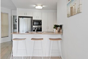 Une cuisine moderne avec des armoires blanches, un four à micro-ondes encastré, un four et un évier en acier inoxydable. On y trouve trois tabourets de bar en bois et une plante en pot sur le comptoir.