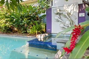 Una piscina con borde de azulejos azules, rodeada de exuberante vegetación y una pared decorativa con un relieve escultórico.
