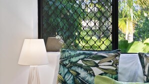 A bedroom with a bed, a nightstand, a lamp, and a window with a view of greenery. - Cairns Hideaway - Pool and Spacious (Cairns)