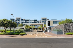 A modern building with multiple floors, a covered entrance, and a landscaped area with trees. - Modern Century City 2 bed Communal Pool Braai (Cape Town)