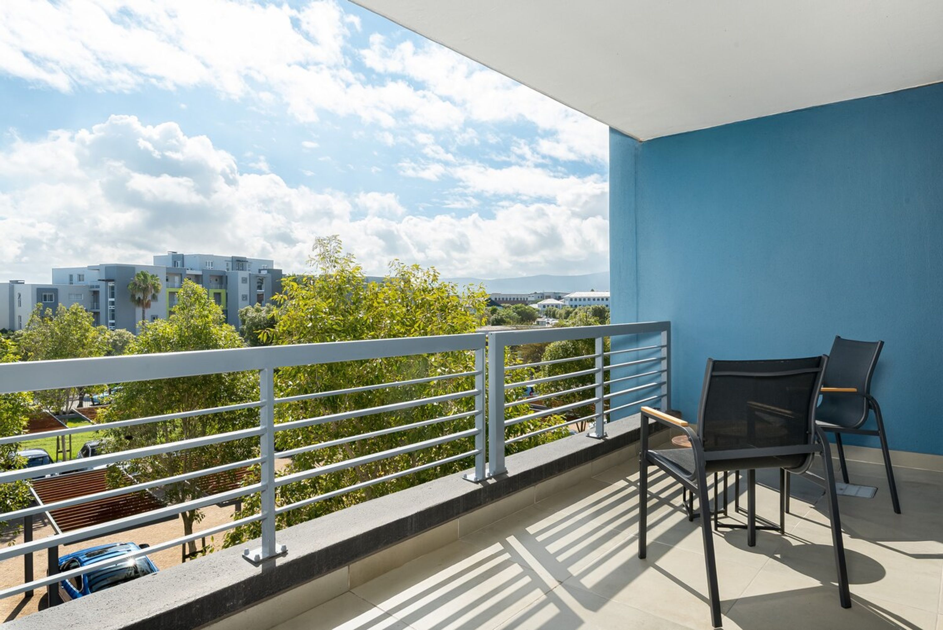 A balcony with a view of buildings and trees, a railing, and two chairs.