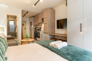 Habitación de hotel pequeña y funcional con cama, una mesita redonda, kitchenette y televisor montado en la pared.
