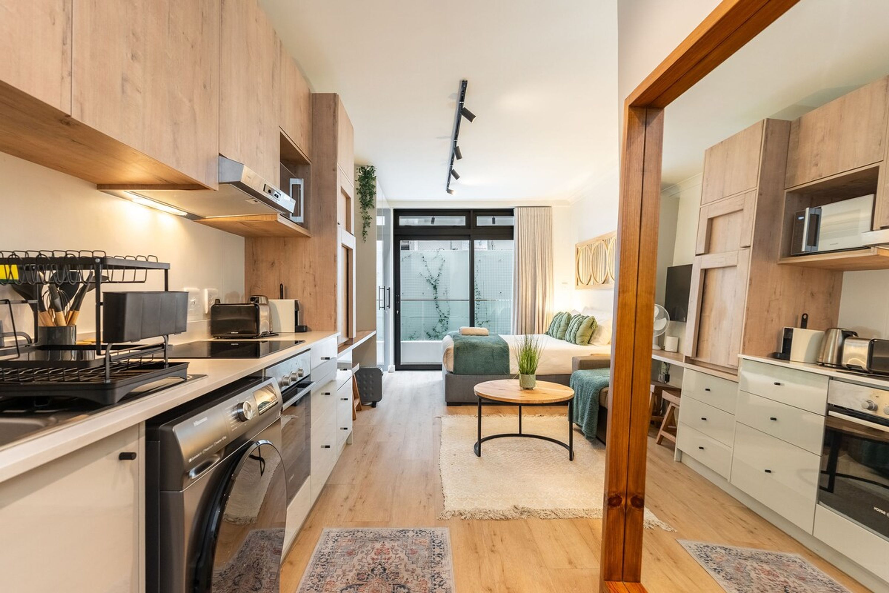 A modern kitchen with integrated appliances, a dining area with a sofa and coffee table, and a view through a glass door to a living room.