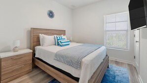 A bedroom with a bed, nightstand, lamp, ceiling fan, window with blinds, and a decorative wall piece. - Over the Dunes at Lost Key Villas (Pensacola)