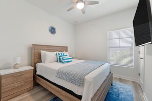 A bedroom with a bed, nightstand, lamp, ceiling fan, window with blinds, and a decorative wall piece. - Over the Dunes at Lost Key Villas (Pensacola)